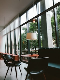 A modern, stylish interior with large floor-to-ceiling windows allowing natural light to flood the space. There is a round wooden table, black chairs, and a comfortable leather sofa. Sheer white curtains hang by the windows, and an elegant floor lamp adds a warm touch. The lush greenery outside is visible through the windows.