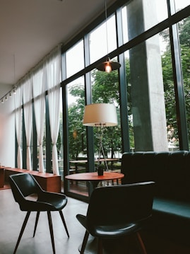 A modern, stylish interior with large floor-to-ceiling windows allowing natural light to flood the space. There is a round wooden table, black chairs, and a comfortable leather sofa. Sheer white curtains hang by the windows, and an elegant floor lamp adds a warm touch. The lush greenery outside is visible through the windows.