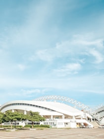 A modern stadium with a curved, white roof and architectural design incorporating metal beams and large windows. The foreground features several green trees and parked cars, all set against a backdrop of a clear blue sky with soft white clouds.