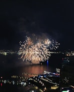 A vibrant display of fireworks lights up the night sky over a body of water, with reflections visible on the surface. The scene includes a waterfront area bustling with people and brightly lit buildings. The city skyline is partially visible in the background, creating a festive atmosphere.