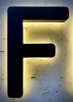 A large black letter 'F' is mounted on a textured, illuminated background that appears to be made of stone or stucco, creating a striking contrast with the surrounding light.