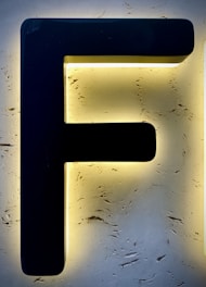 A large black letter 'F' is mounted on a textured, illuminated background that appears to be made of stone or stucco, creating a striking contrast with the surrounding light.