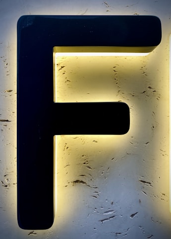 A large black letter 'F' is mounted on a textured, illuminated background that appears to be made of stone or stucco, creating a striking contrast with the surrounding light.
