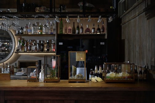 A dimly lit bar features an array of drinks on shelves, with various bottles displayed prominently. Three glass cases sit on the wooden bar counter, containing different beverages and decorative items. Hanging wine glasses and soft lighting add to the cozy ambiance.