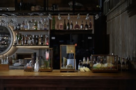 A dimly lit bar features an array of drinks on shelves, with various bottles displayed prominently. Three glass cases sit on the wooden bar counter, containing different beverages and decorative items. Hanging wine glasses and soft lighting add to the cozy ambiance.