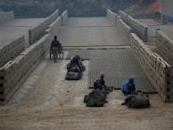 A brick kiln where several workers are molding clay into bricks. Rows of stacked bricks line the sides, and a dirt pathway runs through the middle. Workers are engaged in labor-intensive tasks, with one pushing a wheelbarrow filled with materials.
