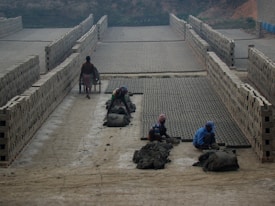 A brick kiln where several workers are molding clay into bricks. Rows of stacked bricks line the sides, and a dirt pathway runs through the middle. Workers are engaged in labor-intensive tasks, with one pushing a wheelbarrow filled with materials.