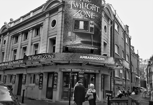 A street view of the Ambassadors Theatre featuring a large 'The Twilight Zone' poster. The theatre entrance is adorned with signage promoting the show. People are walking past and sitting near the entrance. Historic architectural details of the building include arched windows and columns. The scene is in black and white.