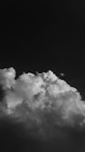 A dramatic, black and white photograph capturing a large, billowing cloud against a dark sky. The cloud appears dense and textured, occupying the lower portion of the image, with intricate patterns and varying shades of gray.