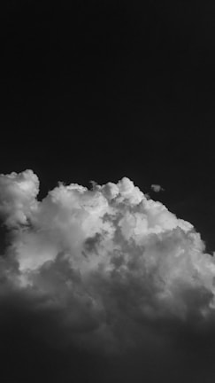 A dramatic, black and white photograph capturing a large, billowing cloud against a dark sky. The cloud appears dense and textured, occupying the lower portion of the image, with intricate patterns and varying shades of gray.
