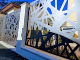 A modern metal fence with an intricate geometric design is prominently featured. The fence has abstract, angular cutouts, creating a pattern that allows glimpses of the background, which includes part of a house and garden. The sky is clear and blue above, adding contrast to the fence's design.