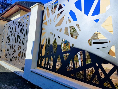 A modern metal fence with an intricate geometric design is prominently featured. The fence has abstract, angular cutouts, creating a pattern that allows glimpses of the background, which includes part of a house and garden. The sky is clear and blue above, adding contrast to the fence's design.