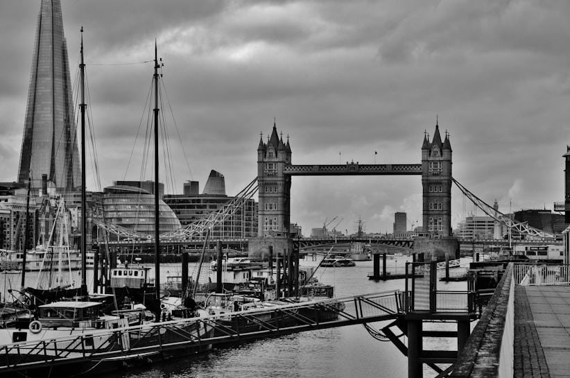 A black and white image of Tower Bridge over the River Thames in London, with The Shard visible to the left. The scene includes various moored boats in the foreground and an overcast sky above, lending a classic and slightly dramatic atmosphere.