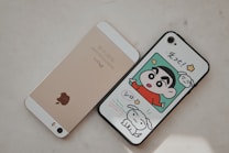 Two smartphones are placed side by side on a light surface. The left phone is gold with a prominent Apple logo on the back. The right phone has a case featuring a colorful cartoon character in a red shirt alongside a white dog, with Japanese text above.