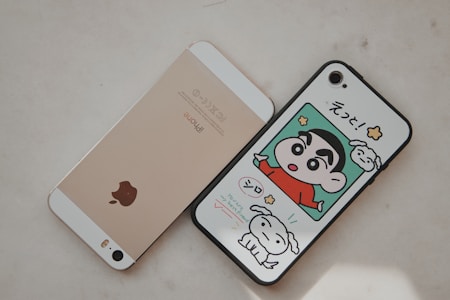 Two smartphones are placed side by side on a light surface. The left phone is gold with a prominent Apple logo on the back. The right phone has a case featuring a colorful cartoon character in a red shirt alongside a white dog, with Japanese text above.