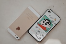 Two smartphones are placed side by side on a light surface. The left phone is gold with a prominent Apple logo on the back. The right phone has a case featuring a colorful cartoon character in a red shirt alongside a white dog, with Japanese text above.