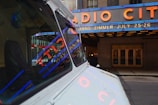 A vintage vehicle is parked on a city street with reflections of bright neon lights in its windows. Large neon signage above an entrance advertises an event featuring Hans Zimmer on July 25-26. The iconic Radio City marquee is prominently displayed in blue and red lighting, set against the stone facade of the building.