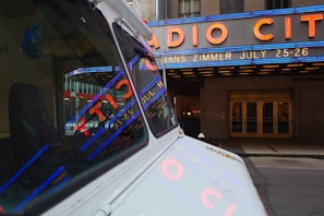 A vintage vehicle is parked on a city street with reflections of bright neon lights in its windows. Large neon signage above an entrance advertises an event featuring Hans Zimmer on July 25-26. The iconic Radio City marquee is prominently displayed in blue and red lighting, set against the stone facade of the building.