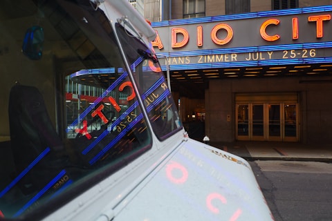 A vintage vehicle is parked on a city street with reflections of bright neon lights in its windows. Large neon signage above an entrance advertises an event featuring Hans Zimmer on July 25-26. The iconic Radio City marquee is prominently displayed in blue and red lighting, set against the stone facade of the building.