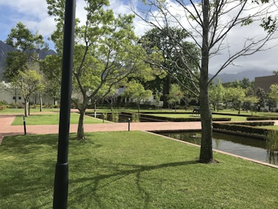 A well-maintained garden features neatly trimmed grass, intersecting brick pathways, and straight lines of small hedges. Trees are scattered throughout the area, some without leaves while others have a full canopy of green foliage. Water bodies with a reflective surface run parallel to the paths, and the sky above is partly cloudy with mountains in the background.