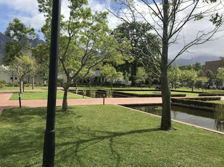 A well-maintained garden features neatly trimmed grass, intersecting brick pathways, and straight lines of small hedges. Trees are scattered throughout the area, some without leaves while others have a full canopy of green foliage. Water bodies with a reflective surface run parallel to the paths, and the sky above is partly cloudy with mountains in the background.