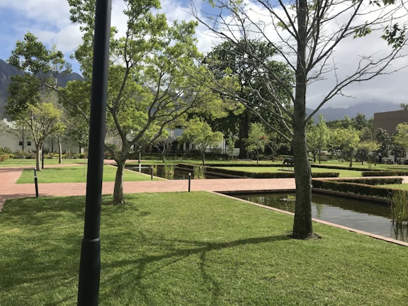 A well-maintained garden features neatly trimmed grass, intersecting brick pathways, and straight lines of small hedges. Trees are scattered throughout the area, some without leaves while others have a full canopy of green foliage. Water bodies with a reflective surface run parallel to the paths, and the sky above is partly cloudy with mountains in the background.