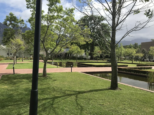 A well-maintained garden features neatly trimmed grass, intersecting brick pathways, and straight lines of small hedges. Trees are scattered throughout the area, some without leaves while others have a full canopy of green foliage. Water bodies with a reflective surface run parallel to the paths, and the sky above is partly cloudy with mountains in the background.