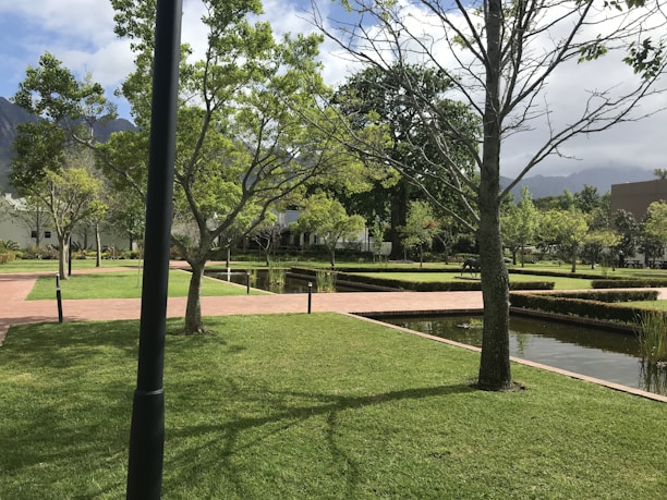 A well-maintained garden features neatly trimmed grass, intersecting brick pathways, and straight lines of small hedges. Trees are scattered throughout the area, some without leaves while others have a full canopy of green foliage. Water bodies with a reflective surface run parallel to the paths, and the sky above is partly cloudy with mountains in the background.