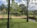 A well-maintained garden features neatly trimmed grass, intersecting brick pathways, and straight lines of small hedges. Trees are scattered throughout the area, some without leaves while others have a full canopy of green foliage. Water bodies with a reflective surface run parallel to the paths, and the sky above is partly cloudy with mountains in the background.