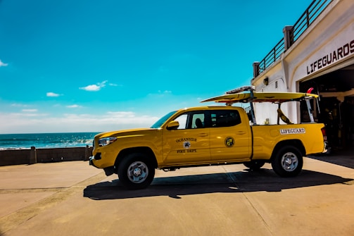 A bright yellow lifeguard truck is parked next to a lifeguard station near the ocean. The truck features lifeguard and fire department insignias and carries surfboards on its rack. The ocean is visible in the background under a wide expanse of blue sky.
