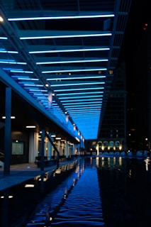 A modern outdoor space is illuminated by rows of vibrant blue lights overhead, creating a striking geometric pattern. Below, a shallow pool of water reflects the lights, adding depth and symmetry to the scene. Buildings can be seen in the background, adding to the urban atmosphere.