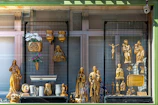A storefront display featuring various golden religious statues and decorative items. The display includes figures of Christian saints and the crucifix, with intricate gold detailing. Floral arrangements in vases and plaques are also part of the presentation.
