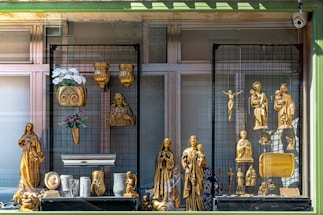 A storefront display featuring various golden religious statues and decorative items. The display includes figures of Christian saints and the crucifix, with intricate gold detailing. Floral arrangements in vases and plaques are also part of the presentation.