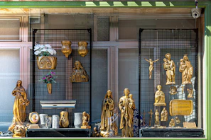 A storefront display featuring various golden religious statues and decorative items. The display includes figures of Christian saints and the crucifix, with intricate gold detailing. Floral arrangements in vases and plaques are also part of the presentation.