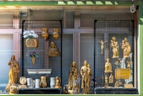 A storefront display featuring various golden religious statues and decorative items. The display includes figures of Christian saints and the crucifix, with intricate gold detailing. Floral arrangements in vases and plaques are also part of the presentation.