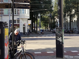 An elderly man is seen adjusting a bicycle on a sidewalk. In the background, there are several street signs indicating directions to various locations. The street is lined with trees and buildings, and there are a few people riding bicycles. A black pole with street art is visible in the foreground. The sun is shining, casting shadows across the scene.