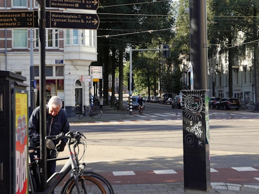 An elderly man is seen adjusting a bicycle on a sidewalk. In the background, there are several street signs indicating directions to various locations. The street is lined with trees and buildings, and there are a few people riding bicycles. A black pole with street art is visible in the foreground. The sun is shining, casting shadows across the scene.