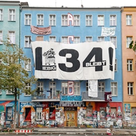A city building facade with a large protest banner reading '34 bleibt.' The building is painted in shades of blue and peach with numerous smaller signs and graffiti covering the lower level. Slogans on various topics including political activism are displayed on the walls. There are trees in front, and apartments with balconies are visible.