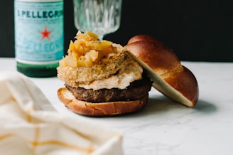 A gourmet burger rests on a marble surface, topped with a generous amount of caramelized onions and a savory sauce. The bun is soft and lightly toasted. In the background, there is a green glass bottle of sparkling water and a partially visible glass, adding a classy, casual dining atmosphere.