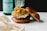 A gourmet burger rests on a marble surface, topped with a generous amount of caramelized onions and a savory sauce. The bun is soft and lightly toasted. In the background, there is a green glass bottle of sparkling water and a partially visible glass, adding a classy, casual dining atmosphere.
