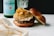 A gourmet burger rests on a marble surface, topped with a generous amount of caramelized onions and a savory sauce. The bun is soft and lightly toasted. In the background, there is a green glass bottle of sparkling water and a partially visible glass, adding a classy, casual dining atmosphere.
