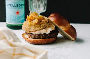 A gourmet burger rests on a marble surface, topped with a generous amount of caramelized onions and a savory sauce. The bun is soft and lightly toasted. In the background, there is a green glass bottle of sparkling water and a partially visible glass, adding a classy, casual dining atmosphere.