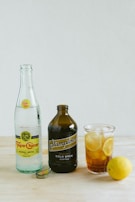 A product display on a light wooden surface featuring a bottle of Topo Chico mineral water, a dark brown bottle of Stumptown cold brew coffee, and a glass filled with iced tea and lemon slices. A lemon sits nearby next to one of the bottle caps.