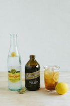 A product display on a light wooden surface featuring a bottle of Topo Chico mineral water, a dark brown bottle of Stumptown cold brew coffee, and a glass filled with iced tea and lemon slices. A lemon sits nearby next to one of the bottle caps.