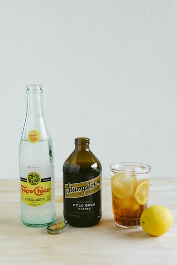 A product display on a light wooden surface featuring a bottle of Topo Chico mineral water, a dark brown bottle of Stumptown cold brew coffee, and a glass filled with iced tea and lemon slices. A lemon sits nearby next to one of the bottle caps.