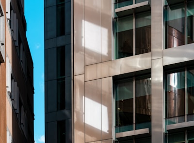 A modern urban architecture scene with close-up views of building facades featuring glass windows and reflective surfaces. The composition includes contrasting materials with sleek metal and textured stone, highlighting the blend of contemporary design elements. Bright sunlight casts distinct shadows and reflections across the surfaces.