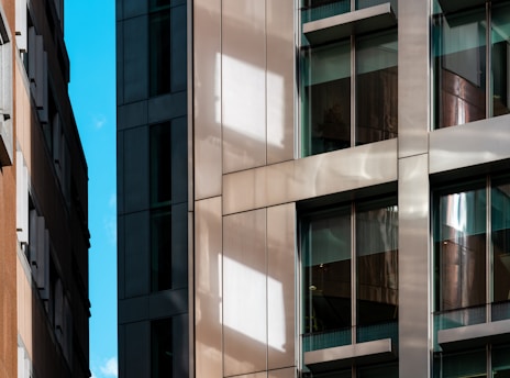 A modern urban architecture scene with close-up views of building facades featuring glass windows and reflective surfaces. The composition includes contrasting materials with sleek metal and textured stone, highlighting the blend of contemporary design elements. Bright sunlight casts distinct shadows and reflections across the surfaces.