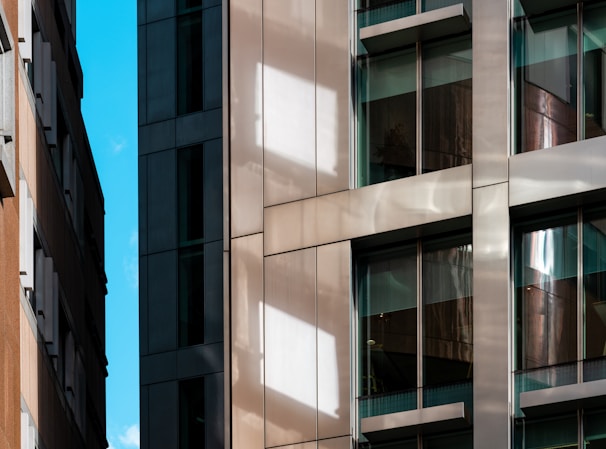 A modern urban architecture scene with close-up views of building facades featuring glass windows and reflective surfaces. The composition includes contrasting materials with sleek metal and textured stone, highlighting the blend of contemporary design elements. Bright sunlight casts distinct shadows and reflections across the surfaces.