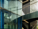 A modern architectural scene featuring a glass facade with visible reflections and metallic frames. The scene includes an internal structure with stairs and metal railings, casting shadows across the glass surface.