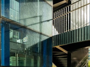 A modern architectural scene featuring a glass facade with visible reflections and metallic frames. The scene includes an internal structure with stairs and metal railings, casting shadows across the glass surface.